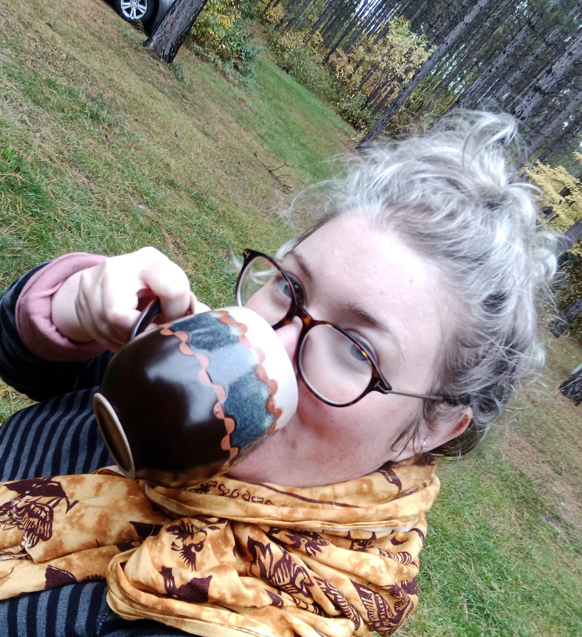 Libby Walkup holding drinking from coffee mug. Pine trees in the background.